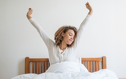 A woman in bed waking up with her arms stretched up high.