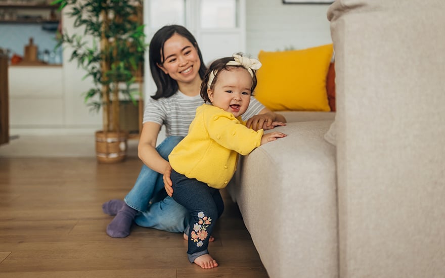 An infant cruises a couch while her mom guides her