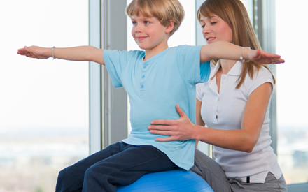 A child sits on a therapy ball while an adult disturbs his balance.