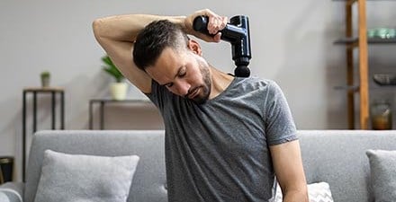 A man using a massage gun on the muscles between his neck and shoulder