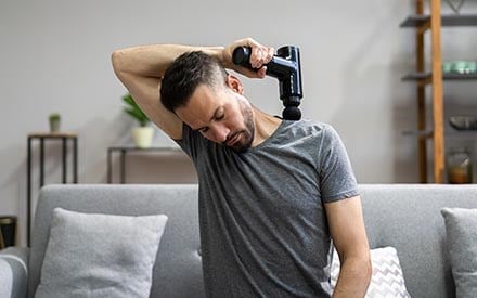 A man using a massage gun on the muscles between his neck and shoulder