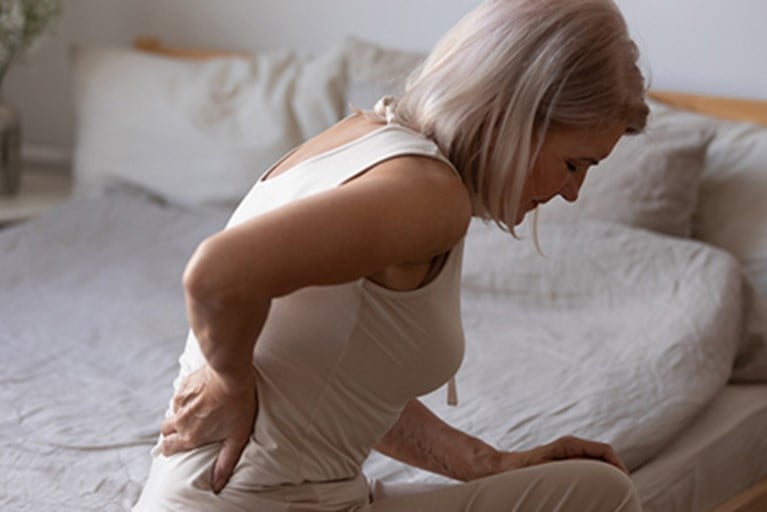A woman sitting on the side of her bed holding her back in pain.