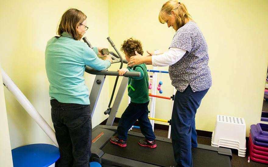 A child walking on a treadmill supported by his physical therapist as mom looks on