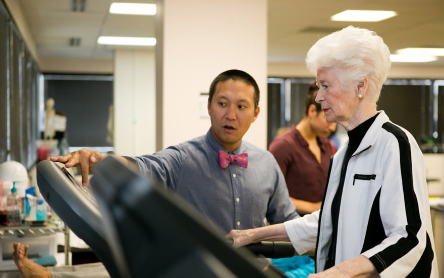 A physical therapist monitors an older adult walking on a treadmill.