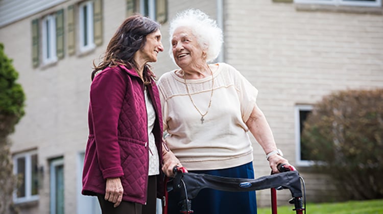 Virginia using a walker outdoors walks smiling toward another women smiling.