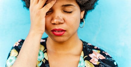 A black female holds her head in pain with her eyes closed.