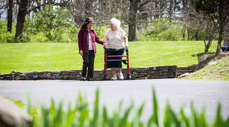 An older adult walking outside with the help of a walker and another person.