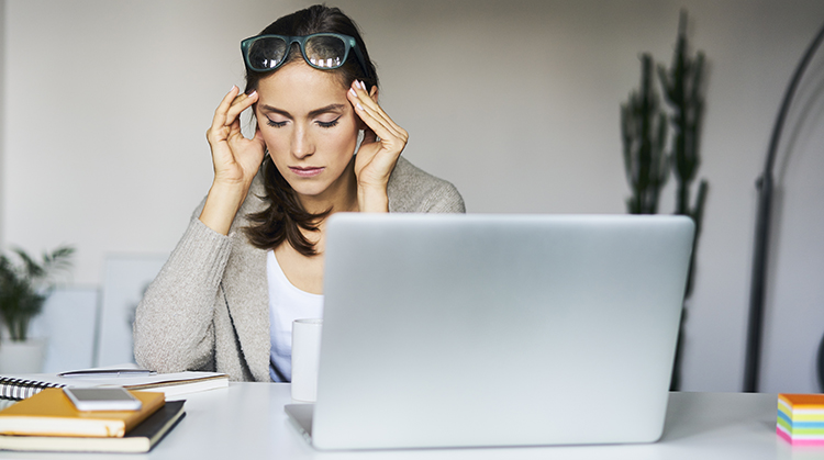 A woman holding her head in pain sitting at a laptop.