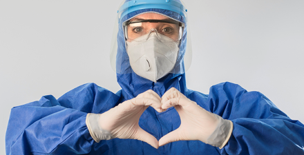 A health care provider dressed in personal protection gear and a mask holding their hands in the shape of a heart.