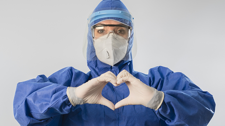 A health care provider dressed in personal protection gear and a mask holding their hands in the shape of a heart.