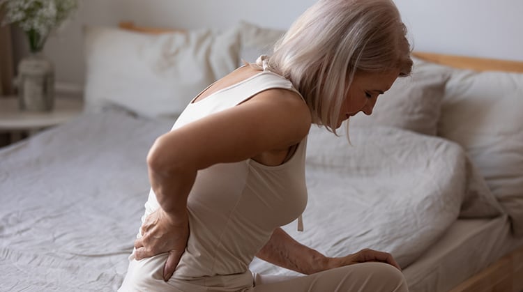 A woman sitting on the side of her bed holding her back in pain.