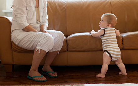 A baby holding the couch walking as mom looks on.