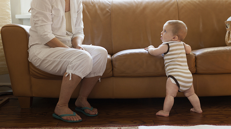 A baby holding the couch walking as mom looks on.