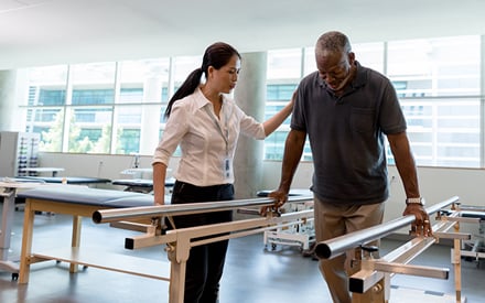 A PT assists a patient using the parallel bars to walk.