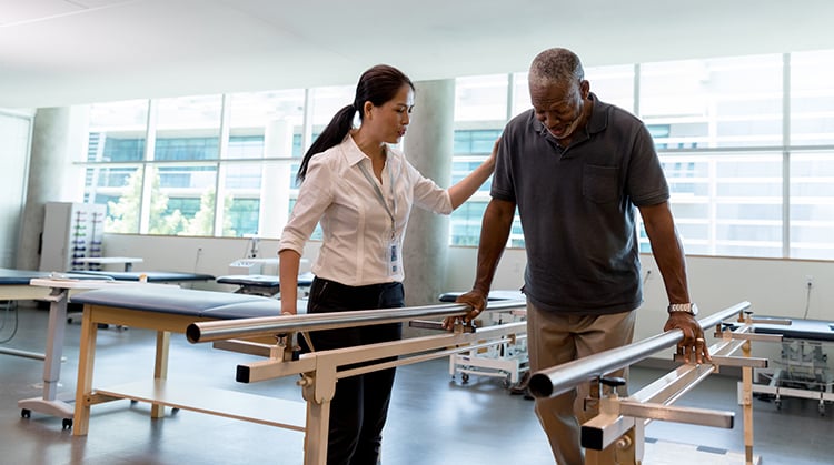 A PT assists a patient using the parallel bars to walk.