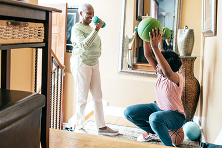 An older adult lifts weights while young family member exercises with a ball.