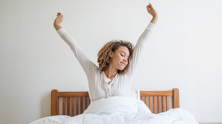 A woman in bed waking up with her arms stretched up high.